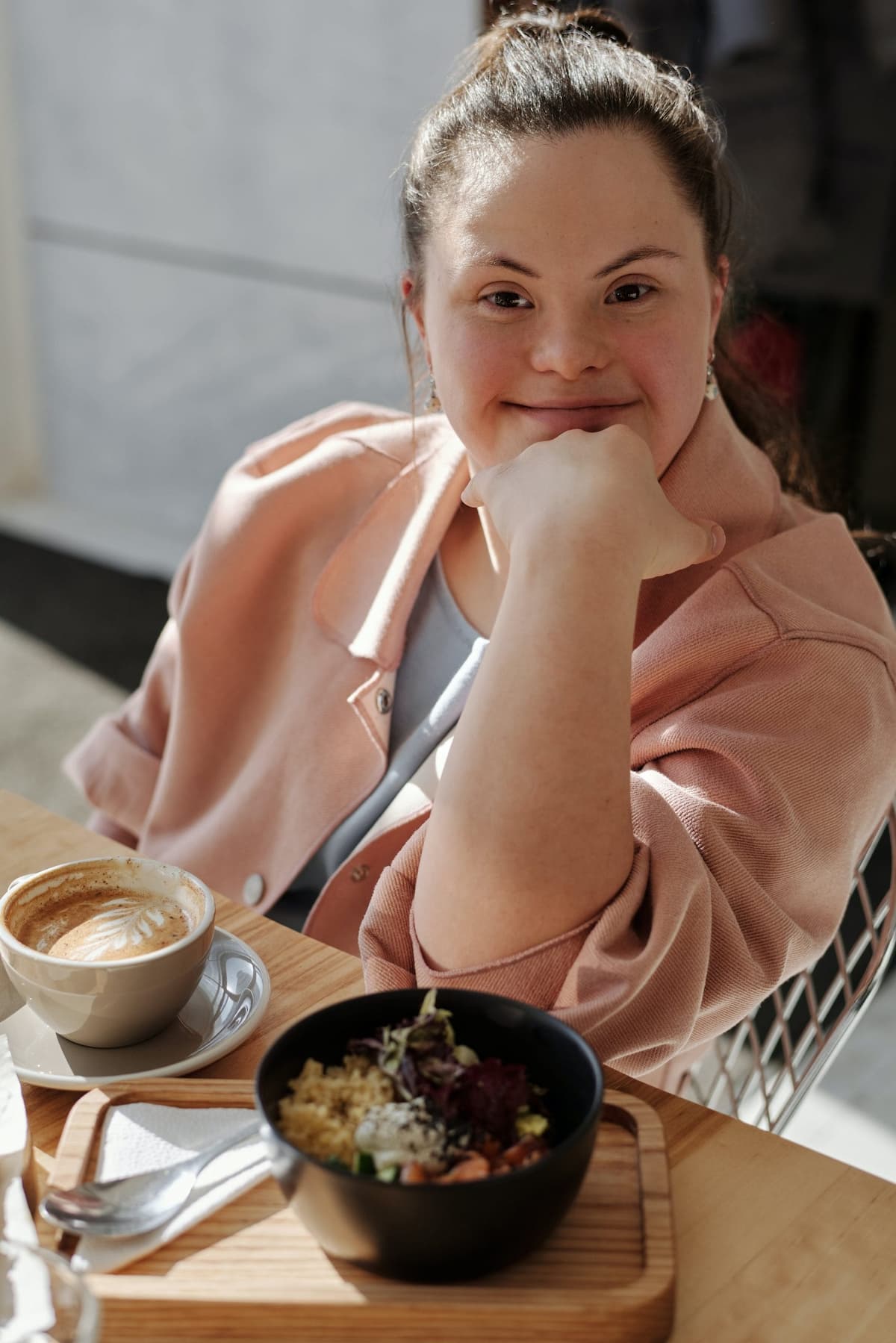 Young woman with down syndrome smiling at the camera. Sitting at a table with a coffee and a meal.